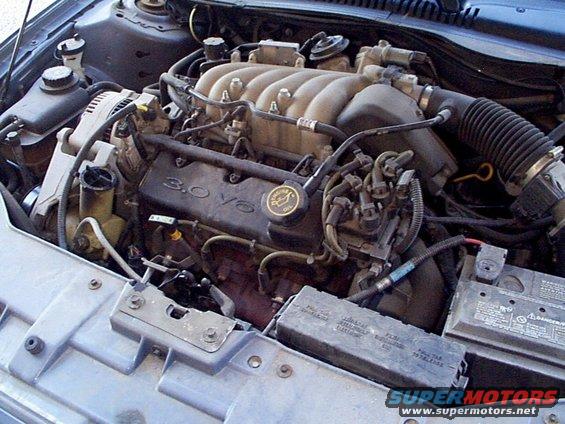 Engine bay of a 1999 Ford Taurus showing the 3.0 V6 engine, battery, and fuse box