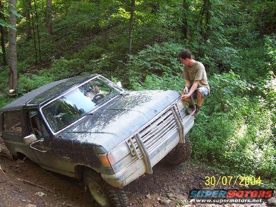 Dark SUV stuck in mud on wooded trail, man on hood