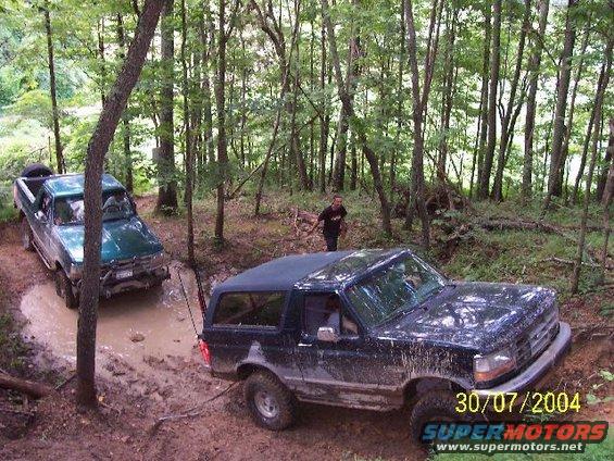 1995 Ford Bronco and a green pickup truck stuck in mud on a forest trail