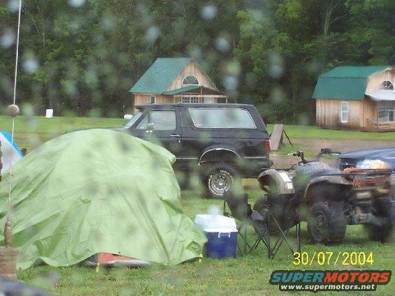 black SUV parked near a tent and an ATV, with two wooden buildings in the background