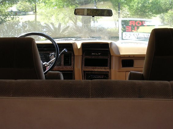 Interior view of a 1985 Ford Bronco XLT, showing dashboard, steering wheel, and seats. A 'For Sale' sign is visible through the windshield