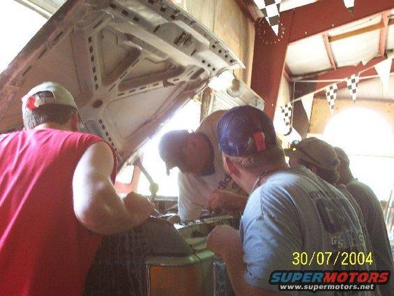 Several guys are gathered around the open hood of a car, looking at the engine. The date 30/07/2004 is visible