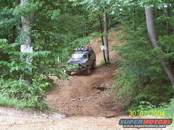 1995 Ford Bronco driving on a dirt trail through trees, equipped with a roof rack and auxiliary lights