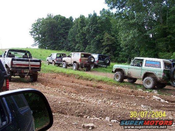 Green 1978 Ford Bronco with white top, off-road tires, and a spare tire on the back, driving on a dirt path