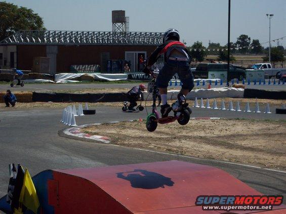 Person on a scooter jumps a ramp at a race track. Another person on a scooter is in the background. Text on bottom right reads SUPERMOTORS