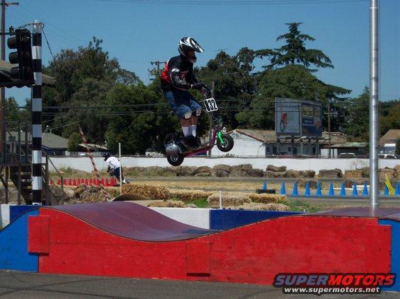 Person on a scooter mid-air over a ramp, wearing a helmet and pads. Number 832 is visible on the scooter