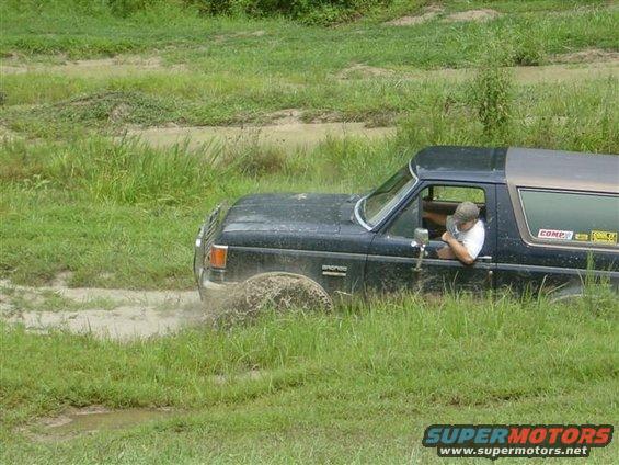 Black Ford Bronco driving through mud and grass, driver visible through the window