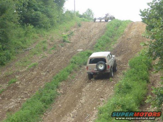 Tan Ford Bronco climbing a steep dirt hill, another vehicle on trailer at top