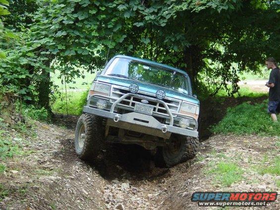 1995 Ford Bronco driving up a steep dirt incline with trees overhead. License plate is visible