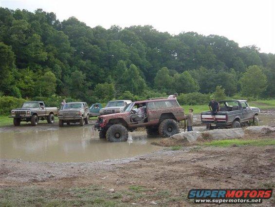Red Ford Bronco stuck in mud, front passenger door open, with other trucks nearby