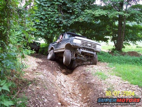 Off-road truck with lifted suspension navigating a muddy ditch, another vehicle visible in background