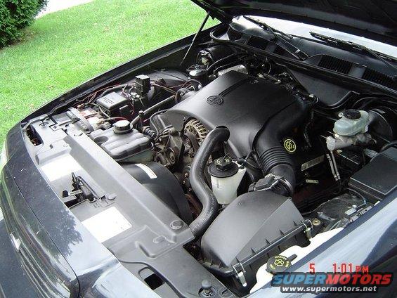 Engine bay of a dark-colored car, hood open. Shows engine, battery, hoses, and air intake