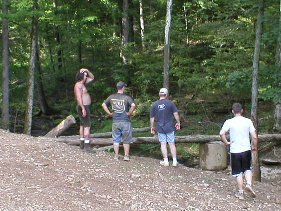 Four men stand in a wooded area near a stream, looking at something off-camera