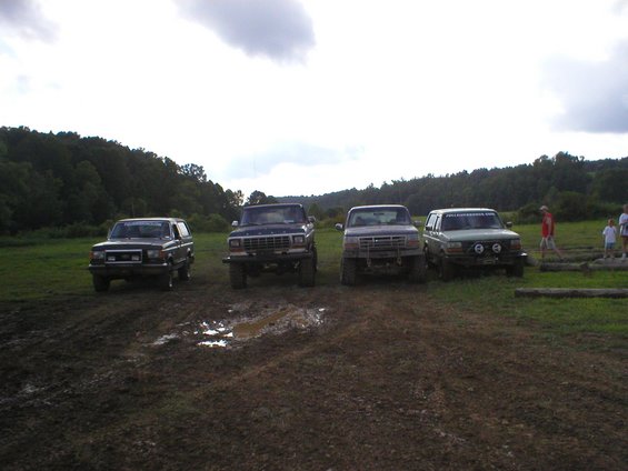 Four trucks parked in mud, with trees and a cloudy sky in the background. Some people are visible to the right