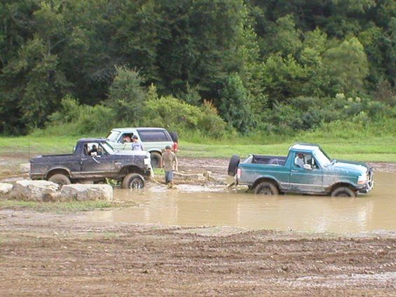 Three off-road trucks stuck in a muddy water pit with trees in the background