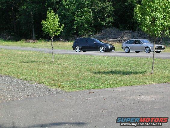 Black hatchback and silver hatchback parked on a road next to a grassy area with trees