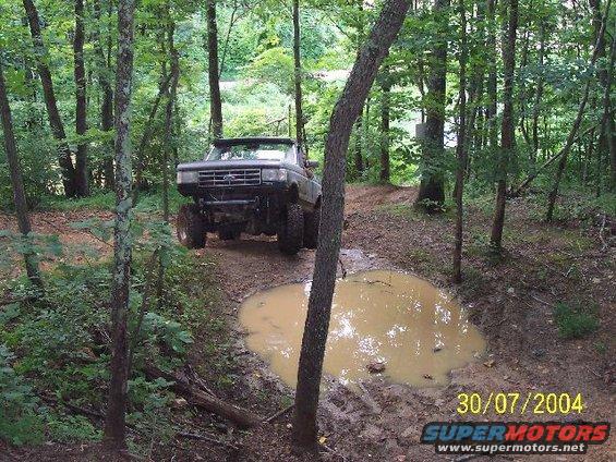 Ford Bronco driving through a muddy forest trail, approaching a large puddle