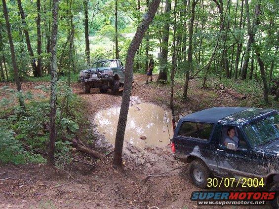 1995 Ford Bronco and a green off-road truck on a muddy path in the woods