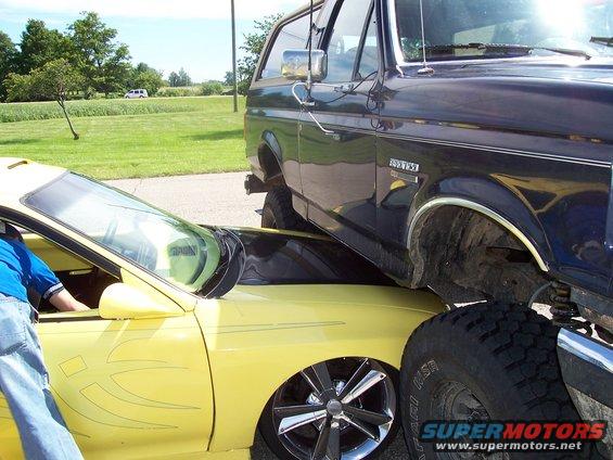 1989 Ford Bronco parked on top of a yellow car, front wheel of Bronco visible