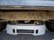 1989 Ford Bronco - The front of a yellow car with custom headlights is tucked under a larger vehicle's chassis.