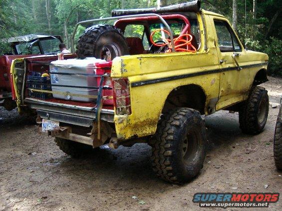 Yellow SUV with spare tire and cooler in the back, parked on dirt road. Another vehicle is partially visible to the left