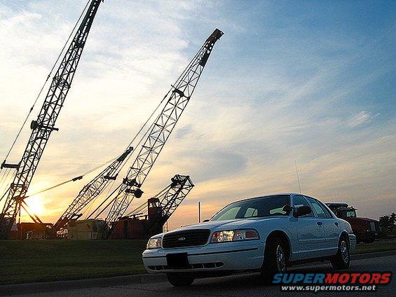 White 2000 Ford Crown Victoria parked on a road with large cranes in the background at sunset