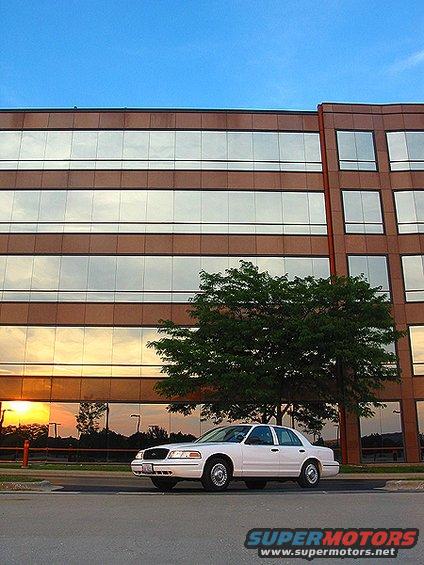 White 2000 Ford Crown Victoria parked on asphalt in front of a modern office building with reflective windows showing a sunset