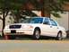 2000 Ford Crown Victoria - A white 2000 Ford Crown Victoria sedan is parked in front of a building with large windows and trees.