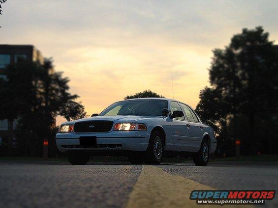 White Ford Crown Victoria parked on road at dusk. Headlights are on