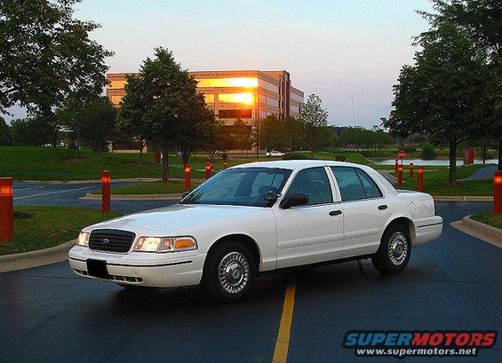 White 2000 Ford Crown Victoria parked on asphalt with yellow lines, sun reflecting off a building