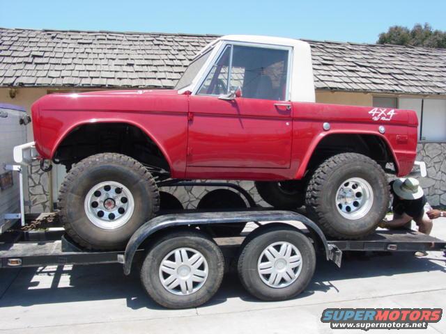 Red first-generation Ford Bronco with white top, lifted on large tires, loaded onto a trailer. A person is working near the rear wheel