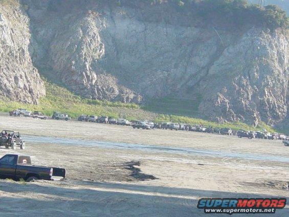 Line of SUVs and trucks parked on a dirt lot with a rocky cliff in the background