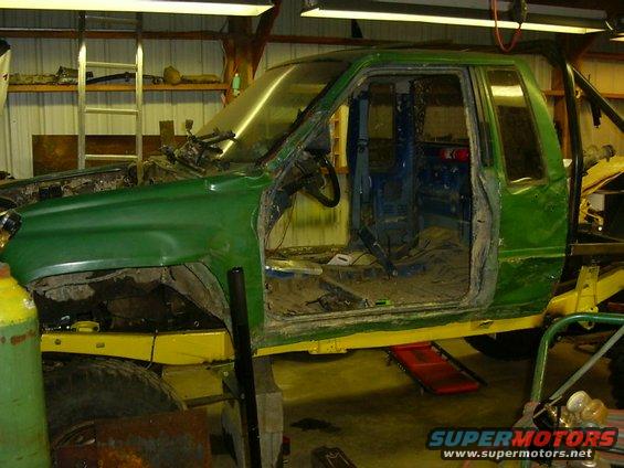 Green pickup truck chassis with yellow frame, missing doors and fenders, in a workshop