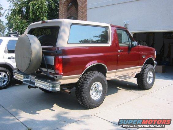 Red Ford Bronco with tan trim and a spare tire on the back, parked in a driveway next to a white SUV