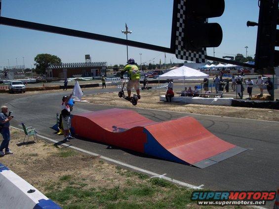 Person on a scooter mid-air over a ramp at a race track. Checkered flag light visible