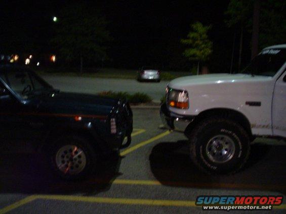 Two trucks parked nose to nose in a parking lot at night. One is dark, the other is white