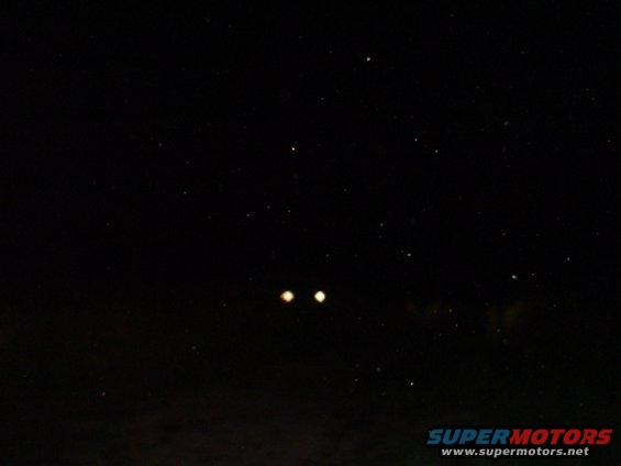 Two glowing headlights in the darkness, with a watermark at the bottom right