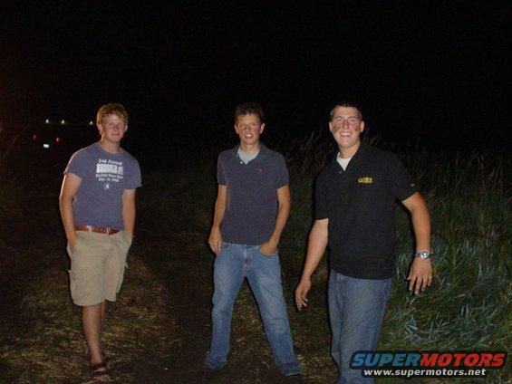 Three young men stand in a grassy area at night, with lights visible in the distance