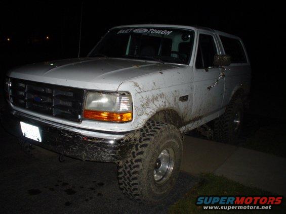 Mud-splattered white Ford Bronco with off-road tires. Banner on windshield reads 'BUILT FORD TOUGH'