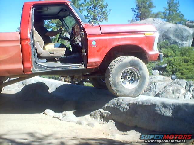 Red pickup truck with open driver's door, driving over rocks. Visible badge on fender