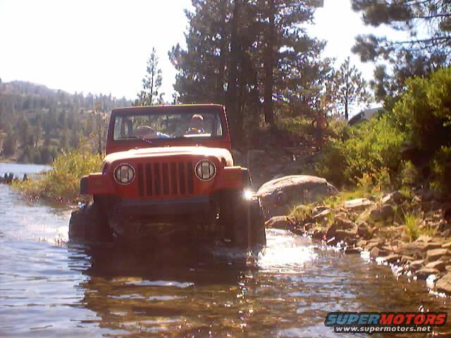 Red Jeep driving through shallow water with trees and rocks in background