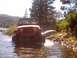 1976 Ford F-100 - A red Jeep navigates through a rocky stream, with trees and a lake visible in the distance.