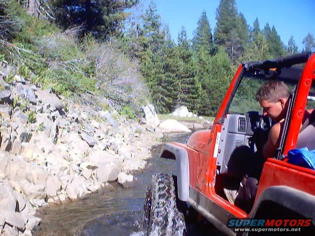 Red Jeep Wrangler driving through a rocky stream with a person in the driver's seat