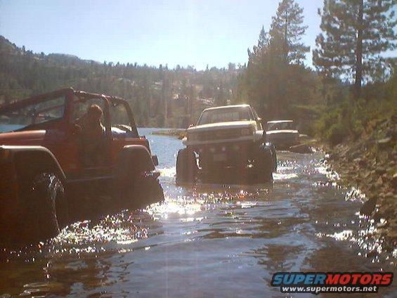 Two Jeeps and a pickup truck driving through a shallow body of water with trees and hills in the background