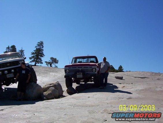 Red 1976 Ford F-100 truck with a winch on the front bumper, parked on a rocky surface next to a white Chevrolet truck