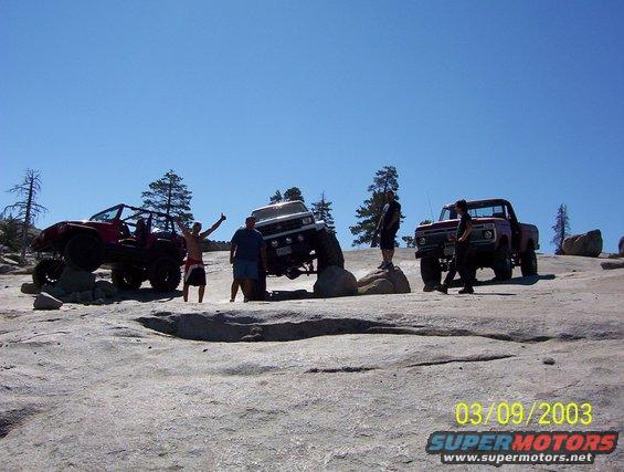 Three off-road vehicles and people on a rocky terrain under a clear blue sky
