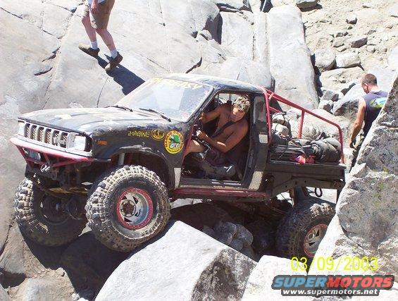 Off-road truck with large tires climbing over rocks. Driver is shirtless