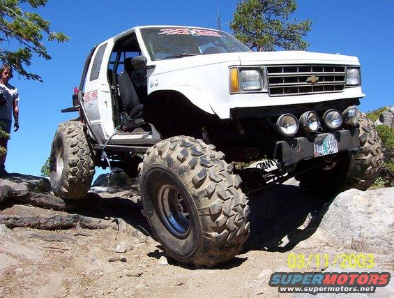 White modified SUV with large tires on rocky terrain, front grille visible