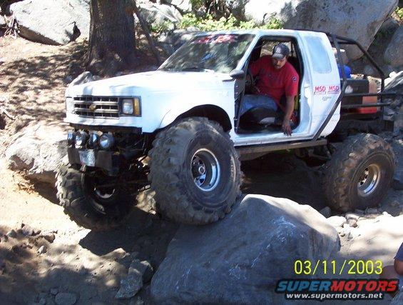 White pickup truck with large tires on rocks, driver in red shirt visible