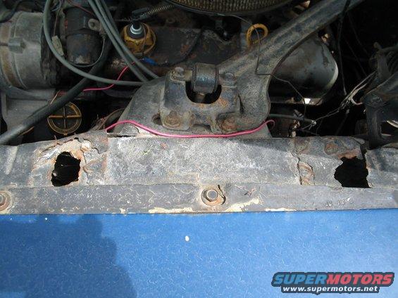 Close-up of a rusted metal car radiator support with holes, showing engine components above and blue body panel below